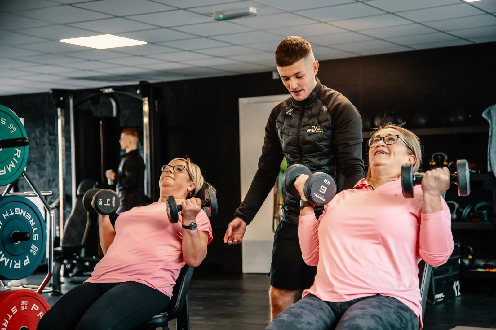 Two members pressing dumbbells side by side while a Level Up Fitness coach helps one of them