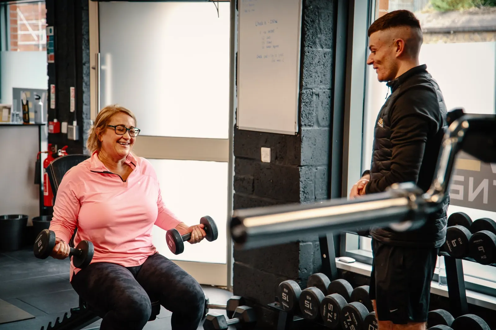 A member smiling as she lifts dumbbells beside a coach at Level Up Fitness in Belfast