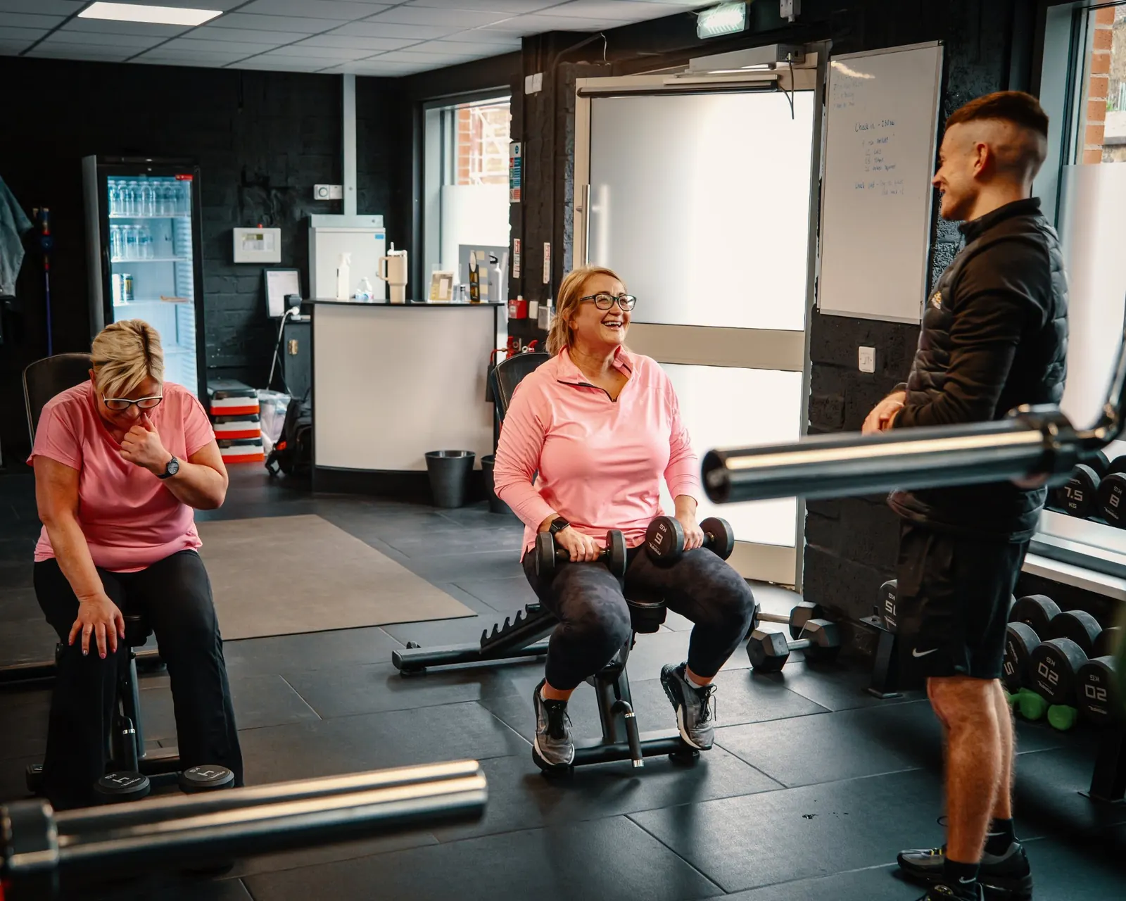 A member laughing between sets of dumbbell work while a Level Up Fitness coach stands with her