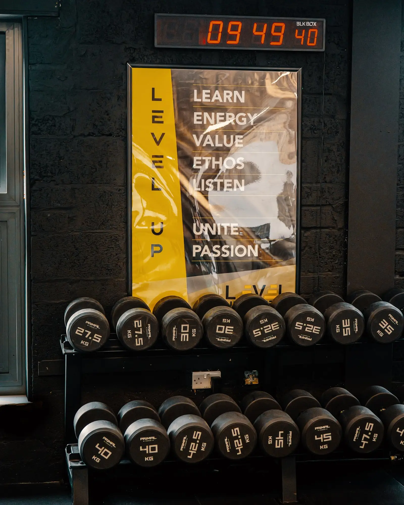 The Level Up poster on the gym wall above the dumbbell rack, spelling out Learn, Energy, Value, Ethos, Listen, Unite, Passion