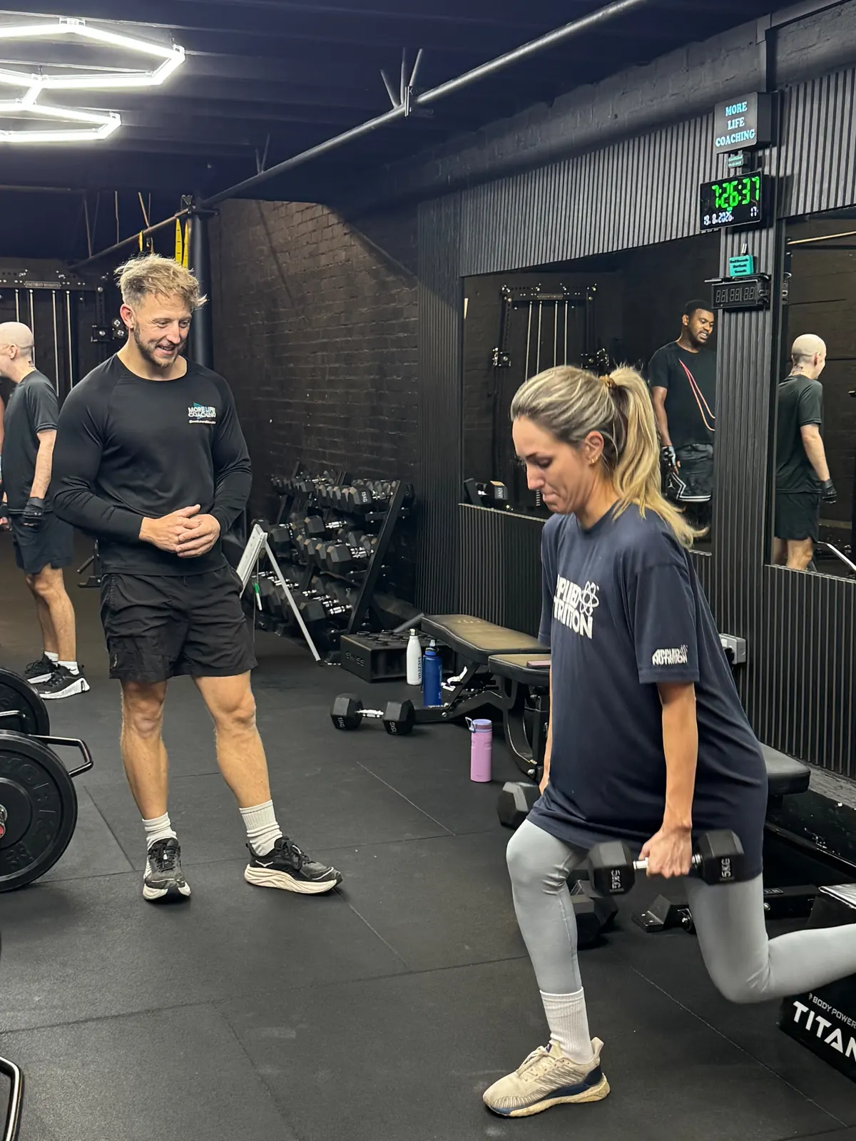 A More Life coach watching a member's form during a dumbbell split squat