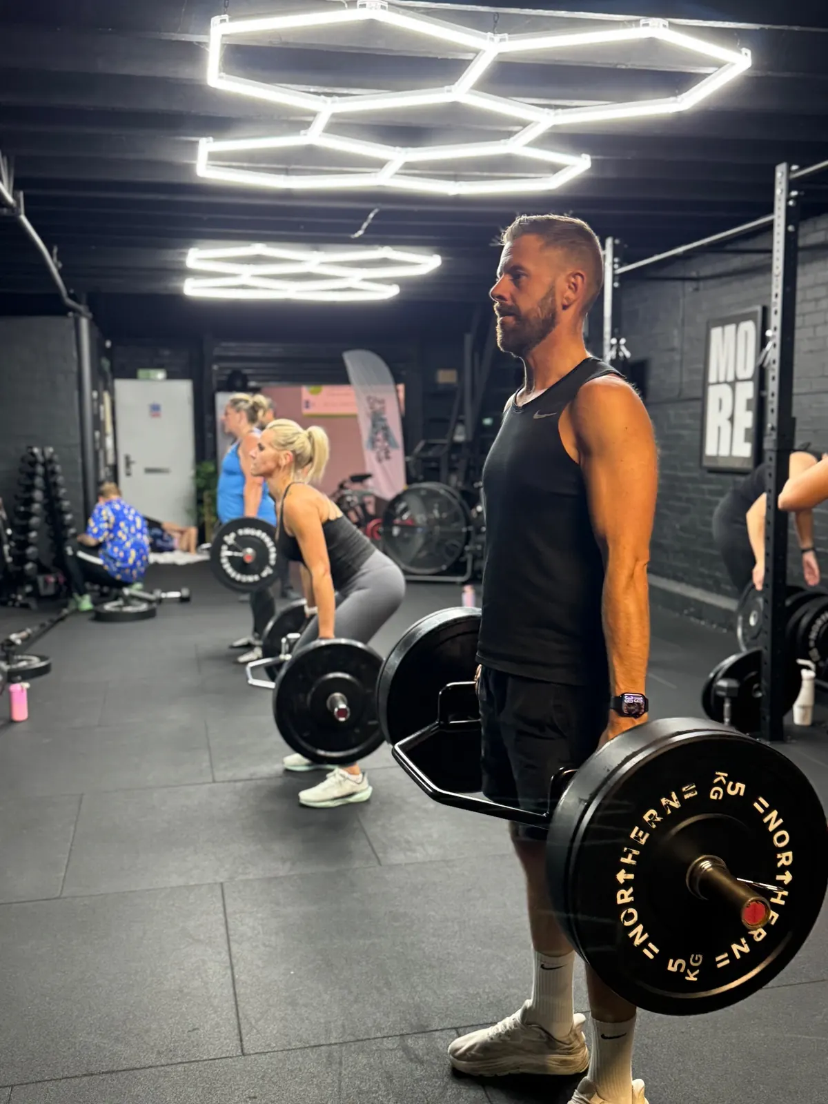 Members lifting barbells in a small group session at More Life Coaching under the hexagon ceiling lights